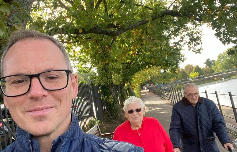 A man wearing glasses and a blue jacket taking a selfie with a woman with short white hair, sunglasses and red jacket and a man wearing a dark jacket using a walker