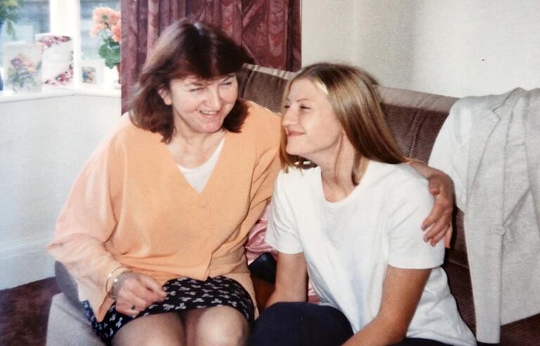 A woman with brown hair wearing an orange jumper and patterned skirt hugging a woman with long blonde hair, white t-shirt and dark trousers while sat on a brown sofa