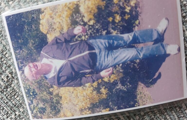 A printed photo of a man stood in front of some flowery bushes