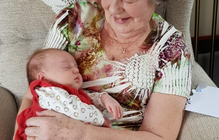 A older woman with short grey hair, wearing a flowery top. She's sat in an armchair and holding a baby
