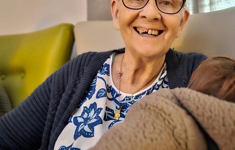 A woman with short grey hair, glasses, patterned top and navy cardigan smiling and sat on a sofa