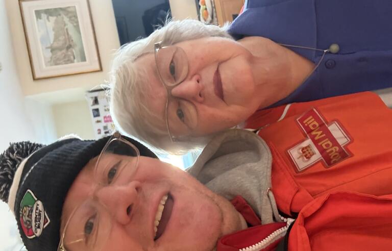A man wearing a Royal Mail jacket, clear glasses and blue bobble hate and a woman with short white hair, clear glasses and blue cardigan