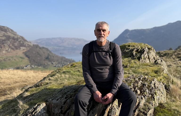 A man wearing a dark top and trousers and glasses with light hair sat on a grassy rock surrounded by hills