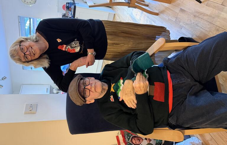 A man sat in a chair wearing a brown hat, glasses, dark jumper and trousers and a woman with a light bob, glasses, dark jumper and gold skirt leaning on the chair