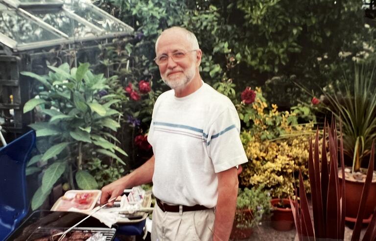 A man with a white beard, white t shirt and beige trousers in a garden