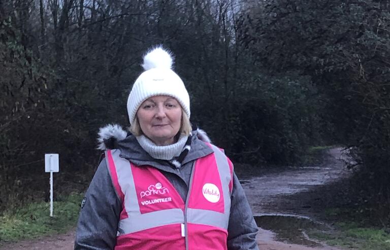 A woman wearing a high viz, hat and coat on a grassy trail