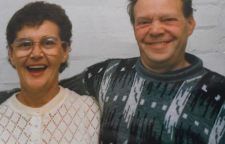 A smiling couple, a lady on the left has short brown hair, glasses and a white cardigan, the man on the right has short brown hair and a green patterned jumper