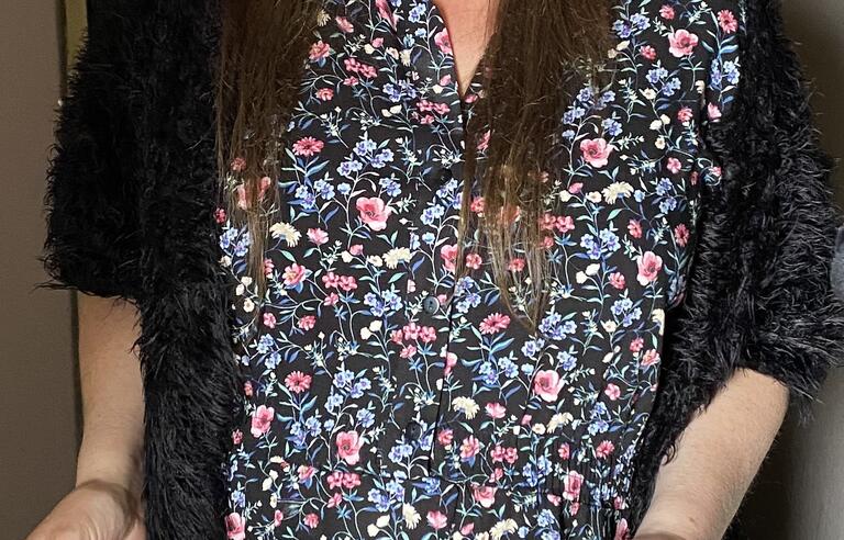 A woman with long brown hair and glasses, long blue patterned dress and dark cardigan
