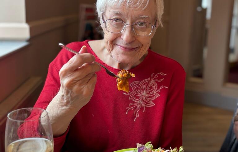 An older woman with short white hair, glasses and wearing a red top eating a lasagna