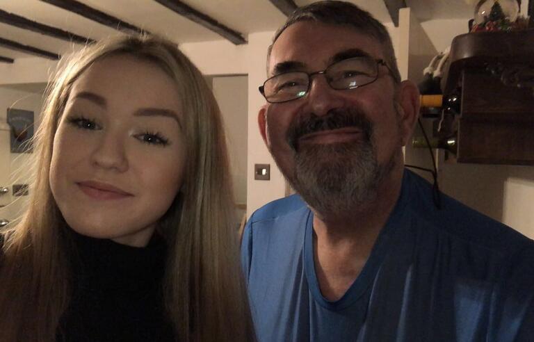 A woman witth long blonde hair taking a selfie with a man with dark hair and a beard wearing glasses and a blue top