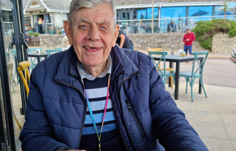 An older man with grey hair, wearing a blue jumper and gilet, drinking a milky coffee