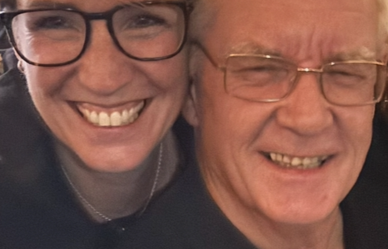 A woman with short blonde hair and glasses hugging an older man with white hair and glasses