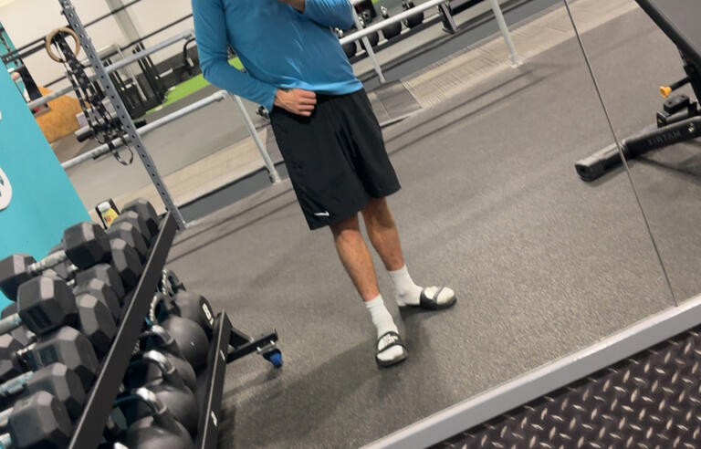 A man wearing a cap, a blue top and shorts taking a selfie in a gym mirror