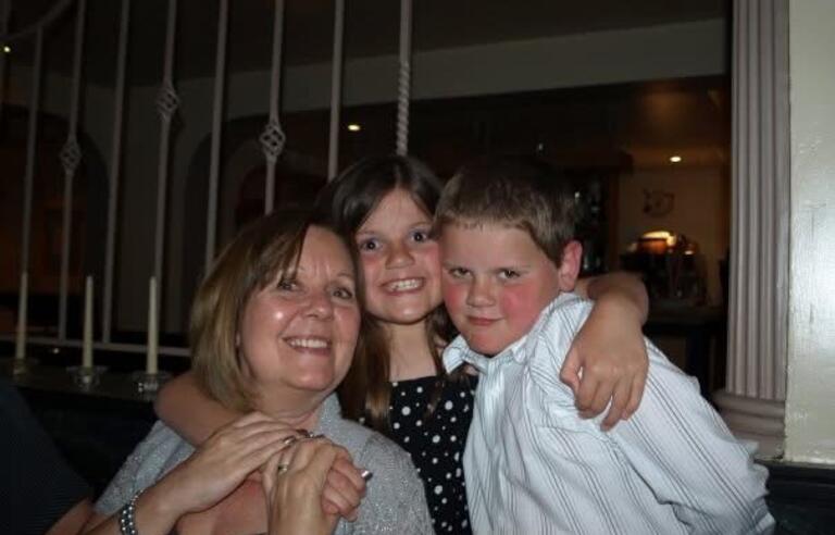 A girl with brown hair and a dark dress hugging a woman with brown hair and grey cardigan and a boy with brown hair and white shirt