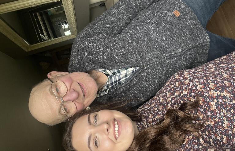 A bald man with glasses wearing a grey cardigan and jeans take a selfie with a woman with long brown hair and a patterned shirt