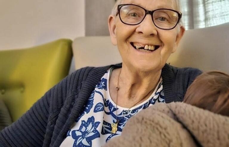 A woman with short blonde hair, glasses, patterned top and blue cardigan holding a brown blanket