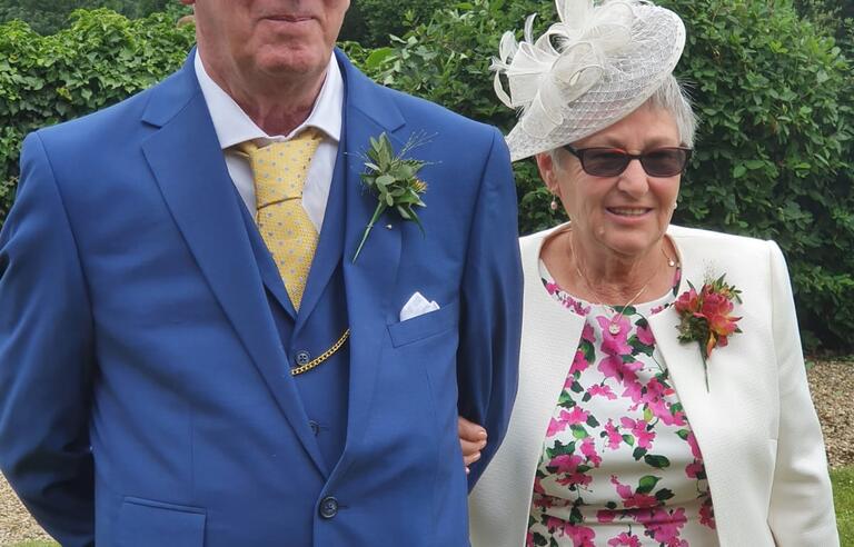A man with white hair and glasses, wearing a blue suit with a white tie and a woman with a white hat and blazer and white and pink floral dress