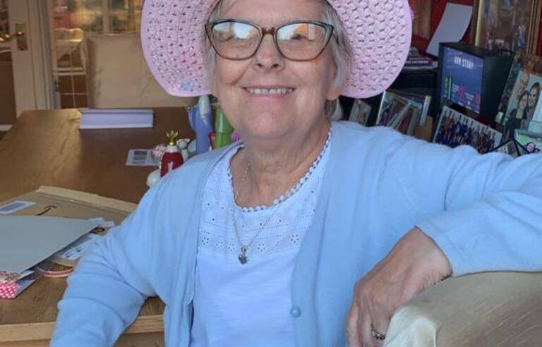 A woman smiling wearing a pink hat, glasses and a light blue top and cardigan